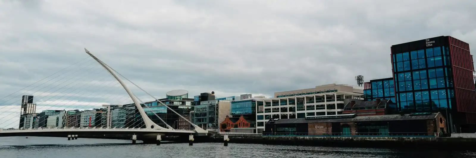 O distrito dos Docklands em Dublin visto do rio Liffey, com a ponte Samuel Beckett à esquerda e os prédios de escritórios envidraçados ao longo do cais.